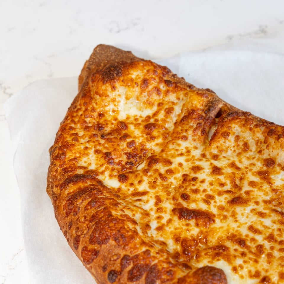 Close-up of a golden-brown, cheesy calzone from Tastivo Pizza on white parchment paper.