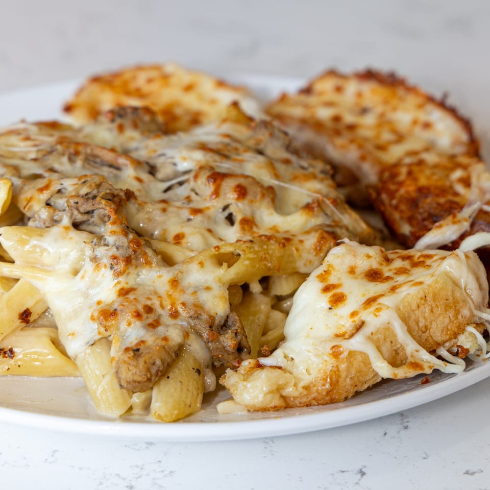 Cheesy baked penne pasta with sausage and two pieces of garlic bread on a white plate.