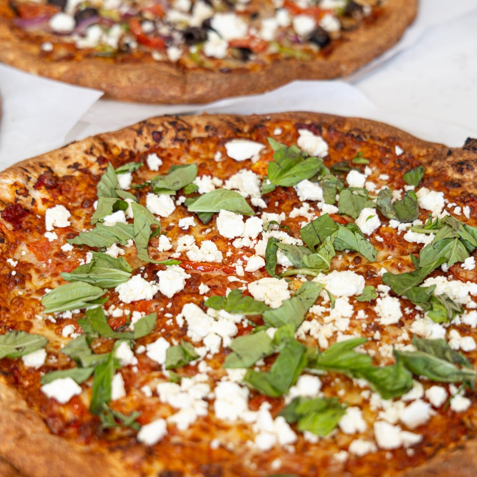 Tastivo Pizza with crumbled feta cheese and fresh basil leaves, with another pizza in the background.