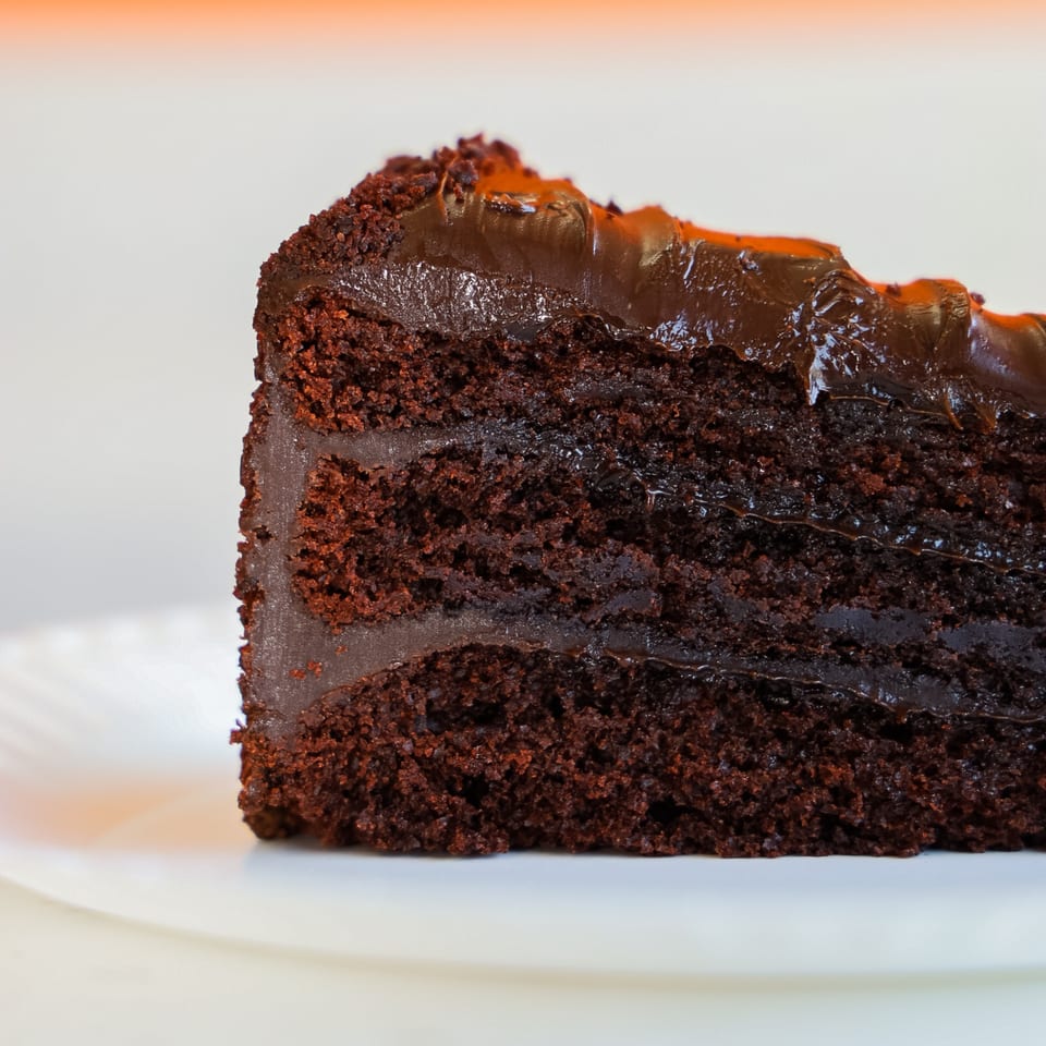 A close-up of a slice of rich, dark chocolate cake with thick chocolate frosting on a white plate.
