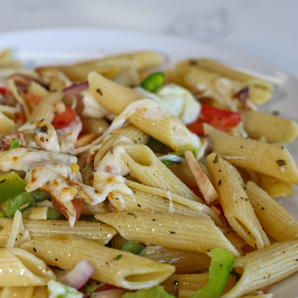 Close-up of penne pasta with melted cheese, green peppers, red tomatoes, and herbs.